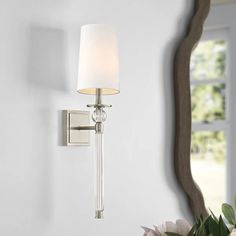 a wall light with a white shade on it next to a flower vase and mirror