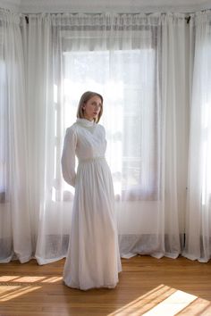 a woman standing in front of a window wearing a long white dress and holding her hands on her hips