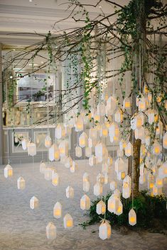 a room filled with lots of white paper lanterns hanging from the ceiling next to a tree