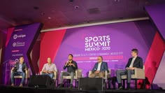 four men sitting on chairs in front of a purple and red backdrop at a sports summit