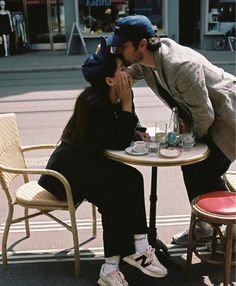 a man and woman sitting at a table with cups on their plates, kissing each other