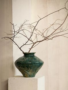 a green vase with branches in it on a white pedestal next to a wall and window