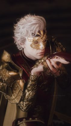 a man with white hair and gold makeup wearing a golden mask, holding his hands to his chest