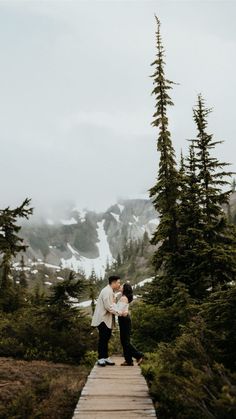 a man and woman standing on a wooden walkway in front of snow covered mountain peaks