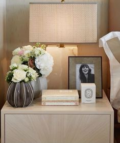 a nightstand with flowers and books on it next to a lamp, photo frame and framed photograph