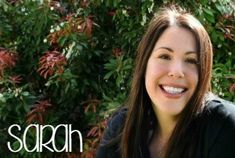 a woman with long brown hair smiling at the camera while sitting in front of some bushes