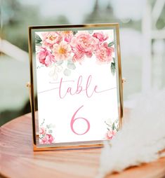 a table number sign with pink flowers and greenery on it sitting on top of a wooden table