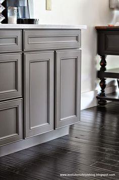 a kitchen with gray cabinets and white counter tops, black wood flooring and a coffee maker on the wall