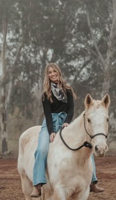 a woman riding on the back of a white horse in a dirt field with trees behind her