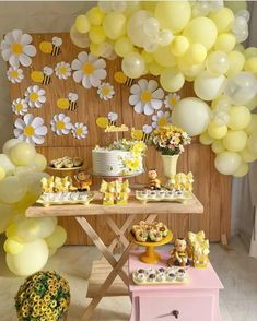 a table topped with lots of yellow and white balloons next to a wall covered in flowers