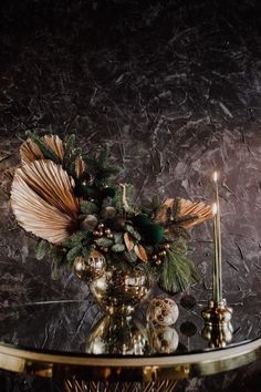 a glass table topped with a gold vase filled with flowers and pineconis next to a candle