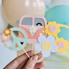 a person holding up some paper flowers and a vw bus cake topper with balloons in the background