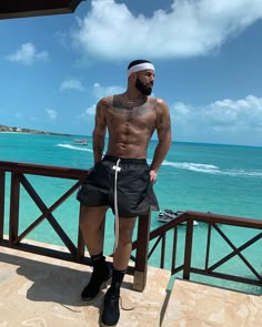 a shirtless man standing on a balcony next to the ocean with his hands in his pockets