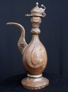 an ornate metal tea pot with a lid on it's side, sitting on a black surface