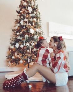 two women sitting on the floor in front of a christmas tree, one is kissing the other's cheek