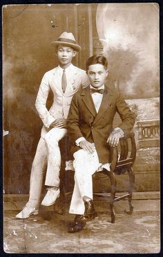 an old black and white photo of two young men in formal wear sitting on a chair