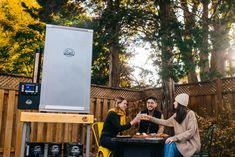 three people sitting at a table with food and drinks in front of an outdoor grill