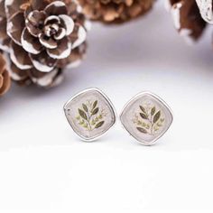 two silver earrings with leaves on them and pine cones in the background, one has been placed