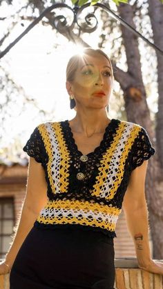 a woman wearing a yellow and black crochet top standing in front of a tree