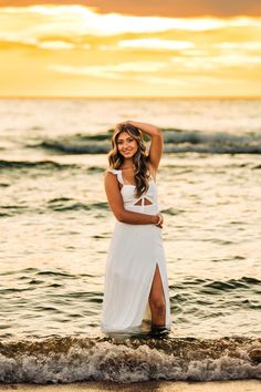 a woman in a white dress standing on the beach with her arms behind her head