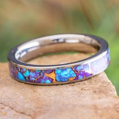 a wedding band with blue and purple flowers inlayed to the side on top of a rock