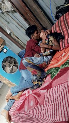 two people sitting on top of a bed with a fan in the air next to them