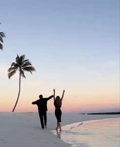 a man and woman walking on the beach with palm trees in the background at sunset