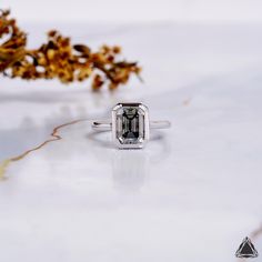 an emerald colored diamond ring sitting on top of a white table next to dried flowers