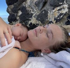 a woman laying on top of a towel next to a baby in her lap with the ocean behind her