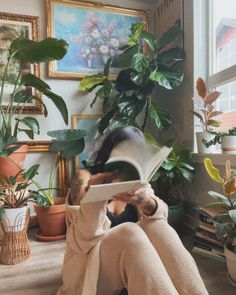 a woman sitting on the floor with her legs crossed reading a book in front of potted plants