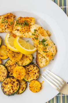 a white plate topped with chicken, zucchini and lemon wedges next to a fork