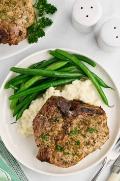 a white plate topped with meat and mashed potatoes next to green beans on a table