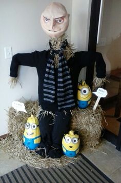 an image of a scarecrow with three minion dolls on hay bales in front of a door