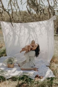 a woman holding a baby in her arms while sitting on a blanket under a tree