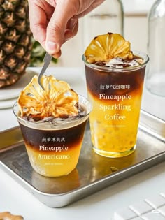 two cups filled with liquid and topped with pineapple on a tray next to another cup