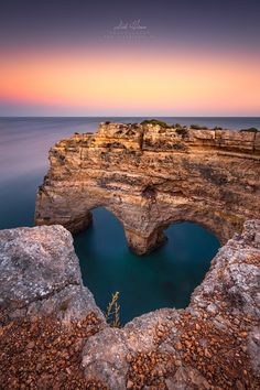 the sun is setting over an ocean with rocks in front of it and a rock arch