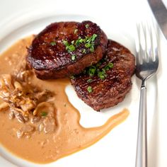 two pieces of steak on a plate with gravy next to a fork and knife