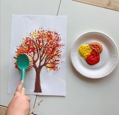 a person holding a green spoon next to a paper plate with an autumn tree on it