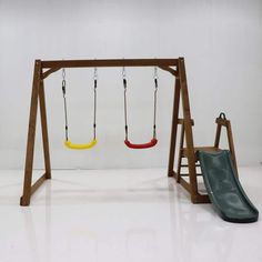 an empty swing set with two swings and a green slide in the foreground, against a white wall