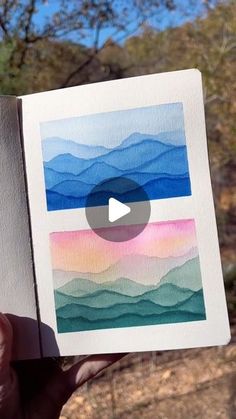 a person holding up a piece of paper with watercolors on it in front of trees