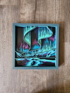 an abstract painting in a blue frame on a wooden surface with the aurora bore visible