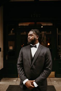 a man wearing a suit and bow tie standing in front of a building with his hands in his pockets