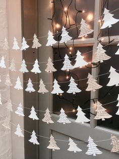 paper christmas trees hanging from strings in front of a window with lights on the windowsill