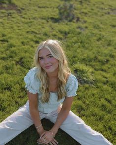 a woman is sitting in the grass with her hands on her knees and looking at the camera