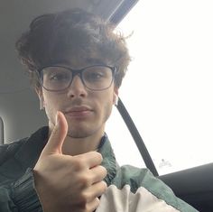 a young man wearing glasses and giving the thumbs up sign in a car with another person behind him