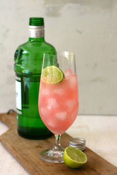 a pink drink in a glass next to a green bottle on a cutting board with limes