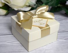 a white box with a gold ribbon and name tag on the lid next to flowers