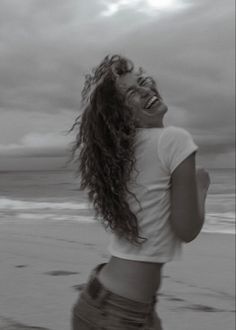 a woman standing on top of a beach next to the ocean with her hair blowing in the wind