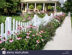 a white picket fence surrounded by flowers and greenery in front of a yellow house