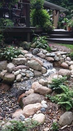 a garden with rocks, plants and water running down the side of it in front of a house